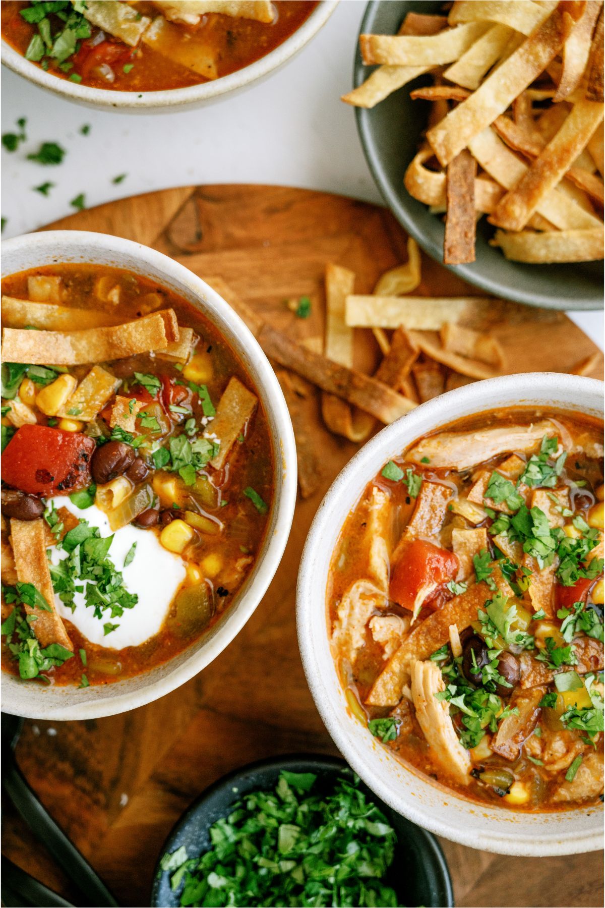 Top view of 2 bowls of Easy Crock Pot Chicken Tortilla Soup, one topped with sour cream. A bowl of tortilla strips and a bowl of chopped cilantro in the background.