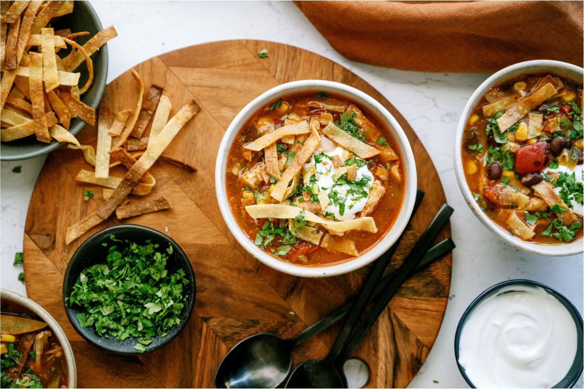 A bowl of Easy Crock Pot Chicken Tortilla Soup topped with tortilla strips, sour cream and chopped cilantro. Another bowl of soup, a bowl of tortilla strips and a small bowl of cilantro in the background.