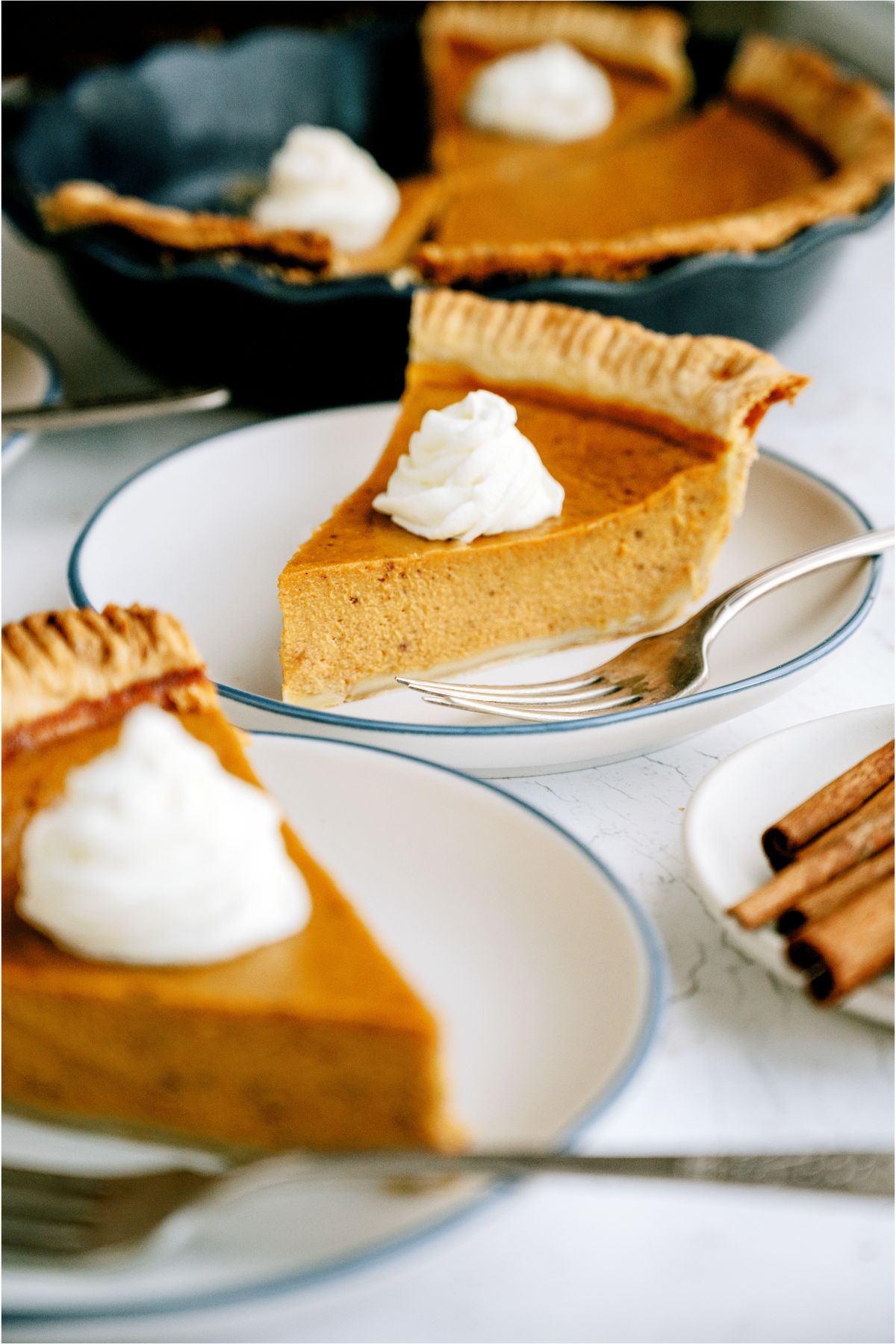 2 plates with slices of Pumpkin Pie topped with whip cream and forks. The remaining pumpkin pie in the pie plate, in the background.