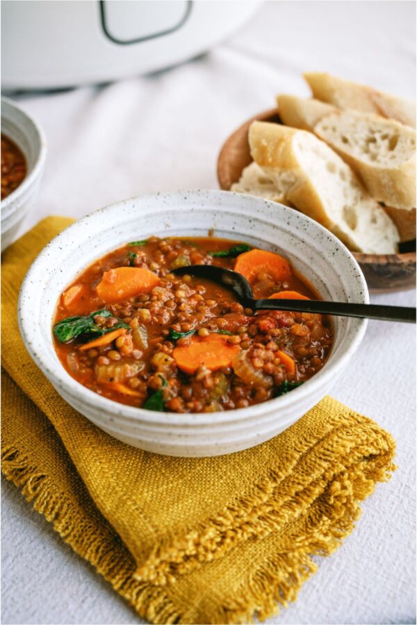 A bowl of Slow Cooker Lentil Soup with a spoon. A side dish of bread and a slow cooker in the background.