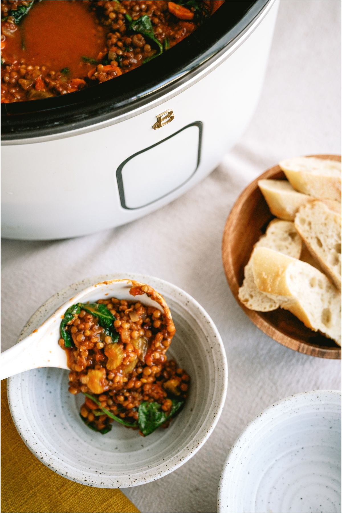 A serving spoon placing a serving of Slow Cooker Lentil Soup into a bowl. With a slow cooker of Slow Cooker Lentil Soup and bowl of bread in the background.