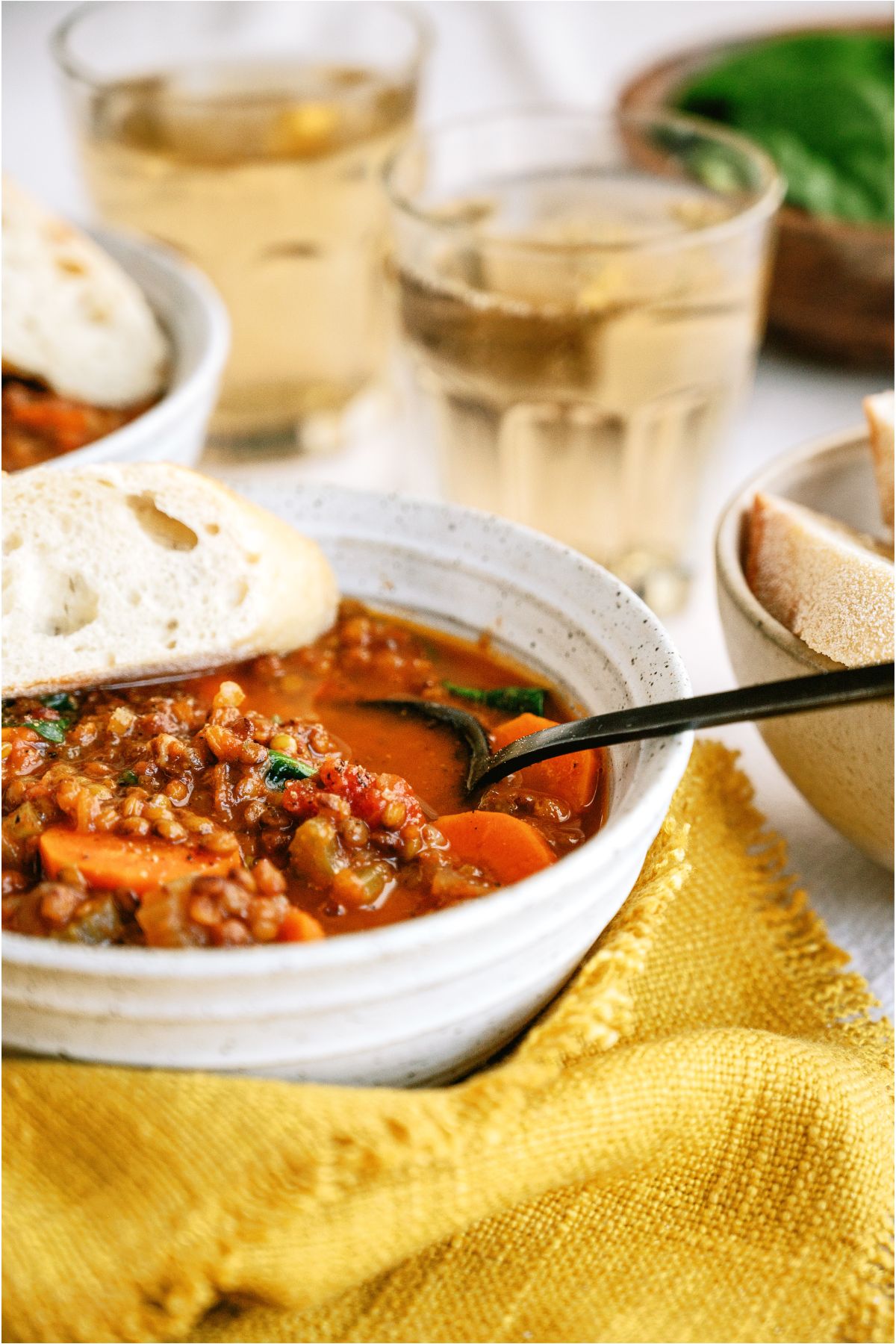 A close up of a bowl of Slow Cooker Lentil Soup with a spoon in it and a piece of bread. With a 2 glasses of water in the background.