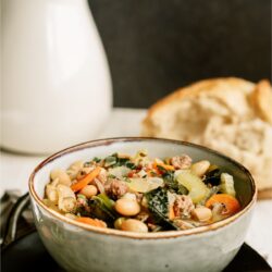 A bowl of Tuscan White Bean Soup with a white pitcher and bread on a plate in the background.