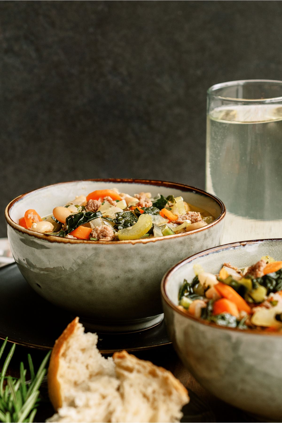 2 bowls of Tuscan White Bean Soup with a slice of bread in front and a glass of water in the background.