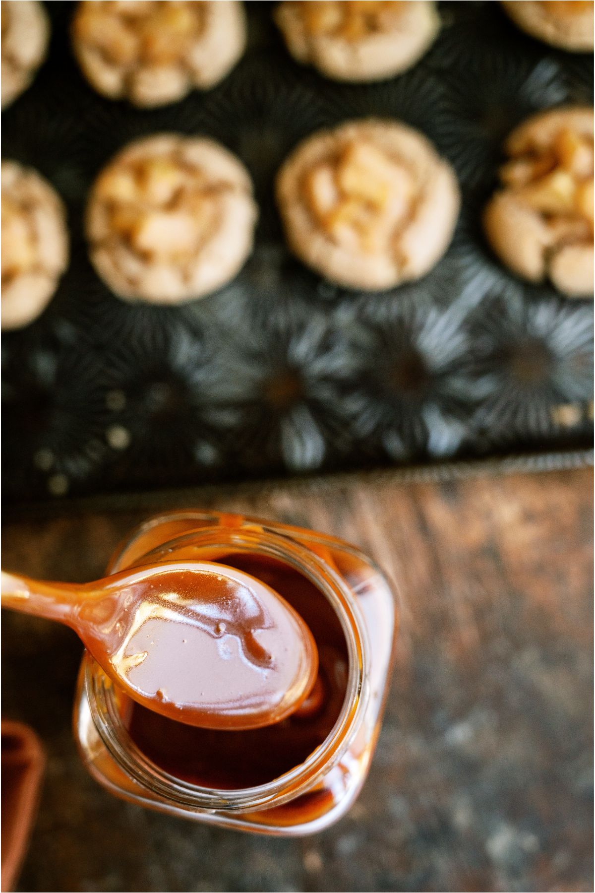 A jar of caramel sauce with a spoon lifting some out. A pan of Apple Pie Cookies in the background.