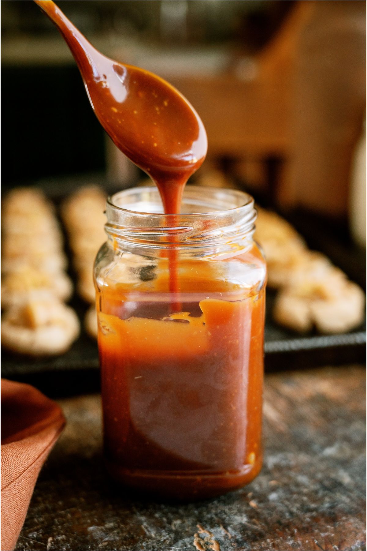 A jar of homemade caramel sauce with a spoon lifting some out. Apple Pie Cookies in the background.