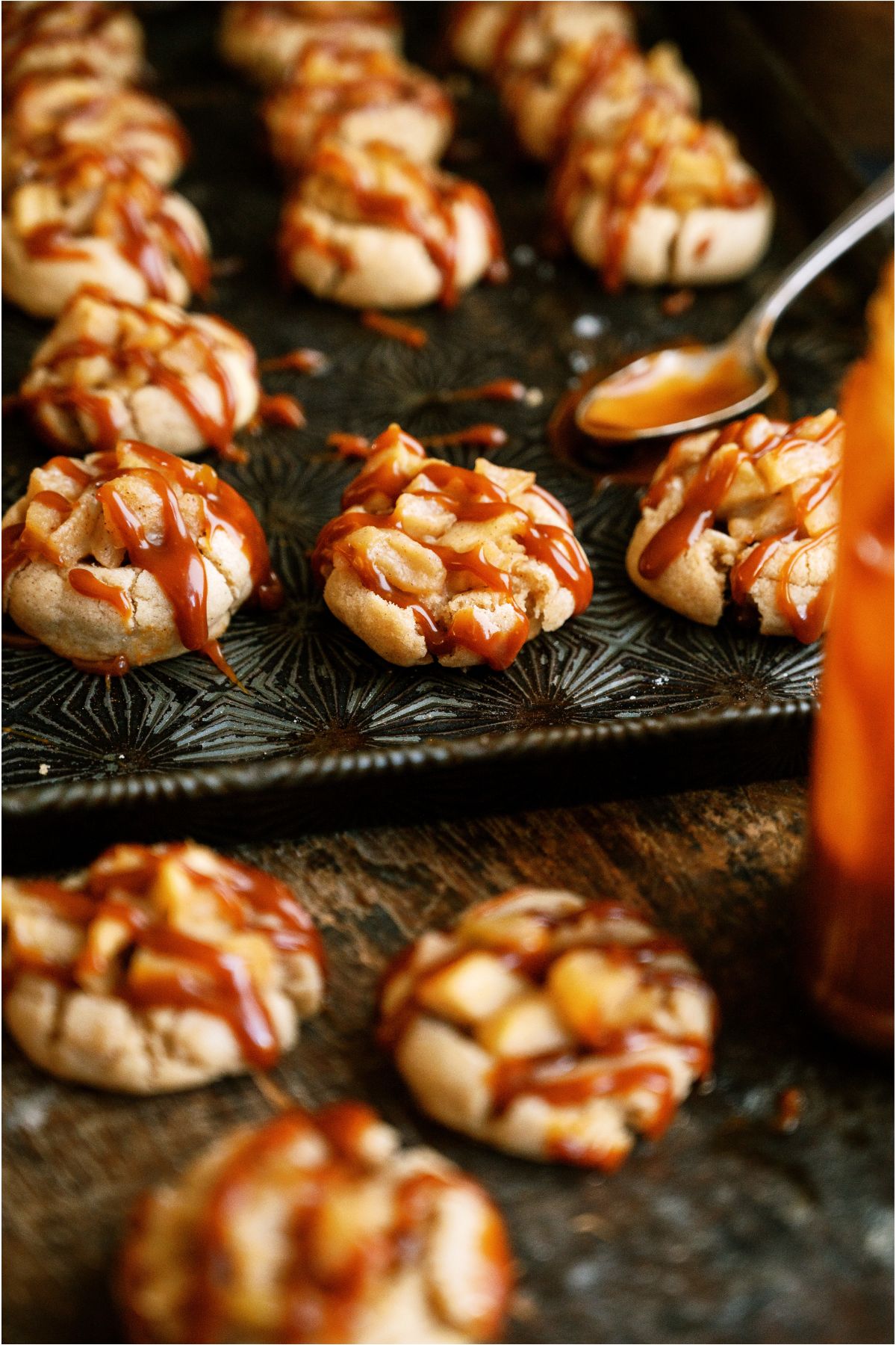 Apple Pie Cookies drizzled with caramel sauce on a pan and on the counter. A spoon with caramel sauce in the background.