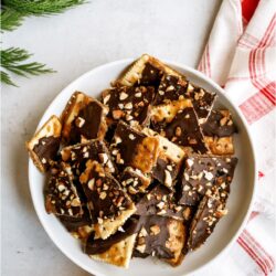 A plate full of broken pieces of Christmas Crack. With a sprig of a pine tree and a Christmas towel underneath the bowl.