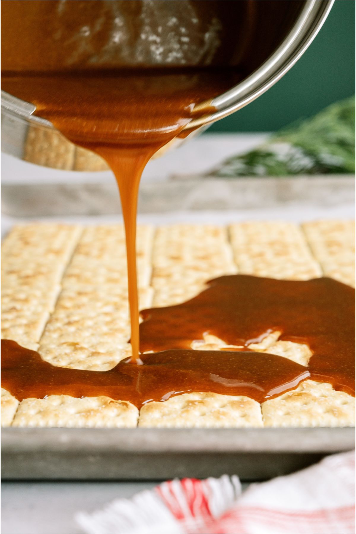 Pouring homemade caramel from saucepan onto sheet pan with lined up saltine crackers.