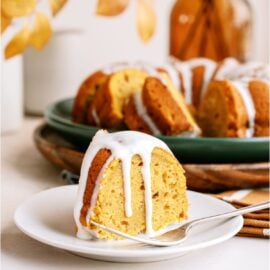 A slice of pumpkin bundt cake with cream cheese glaze on a white plate with the remaining bundt cake with glaze in the background.