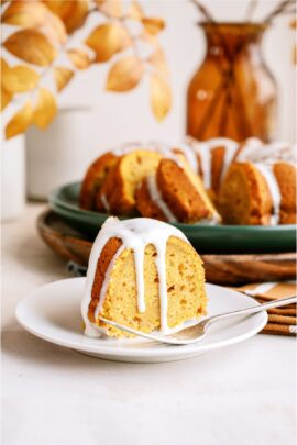 A slice of pumpkin bundt cake with cream cheese glaze on a white plate with the remaining bundt cake with glaze in the background.