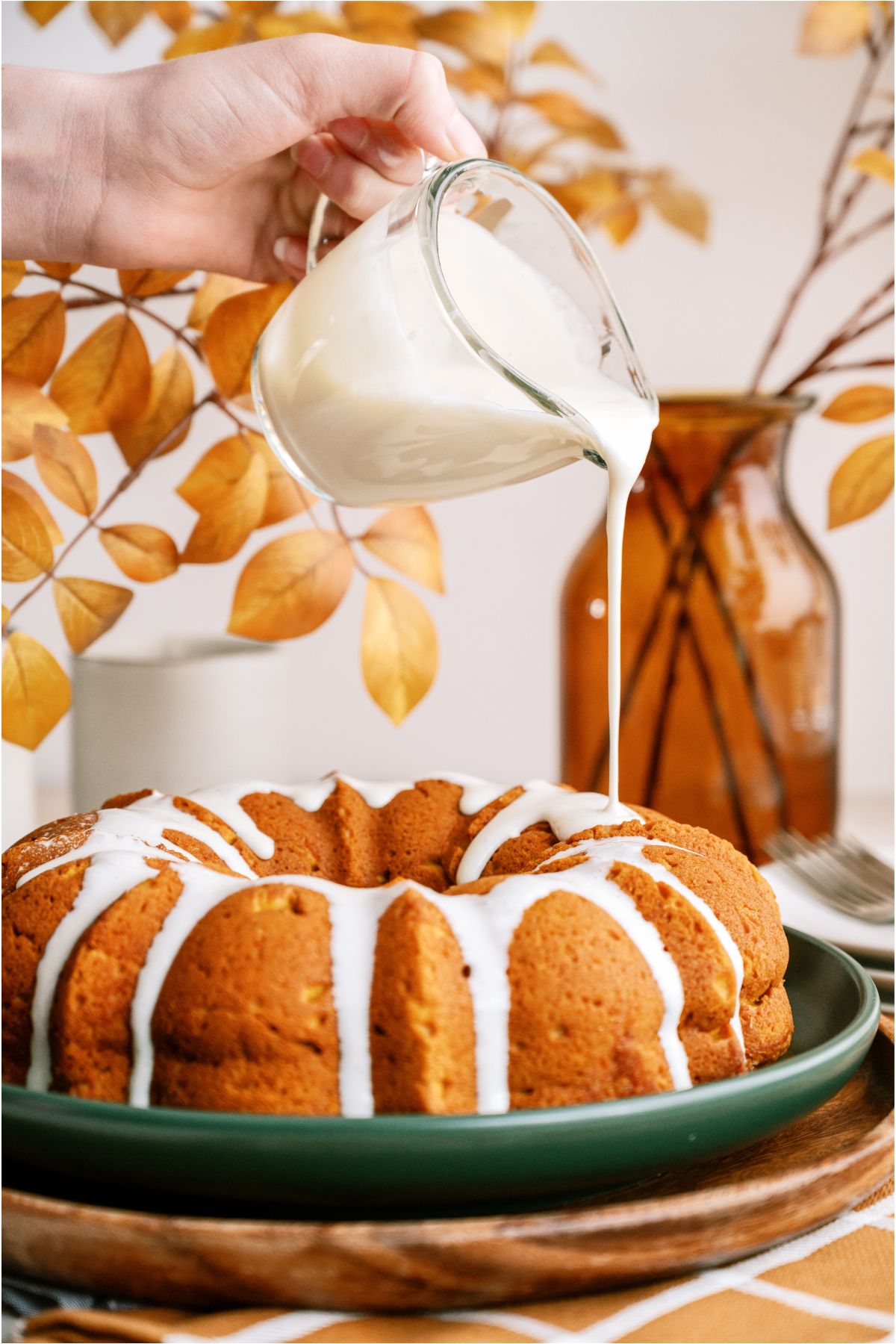 A hand holding a measuring cup pouring cream cheese glaze on top of the Pumpkin Bundt Cake.