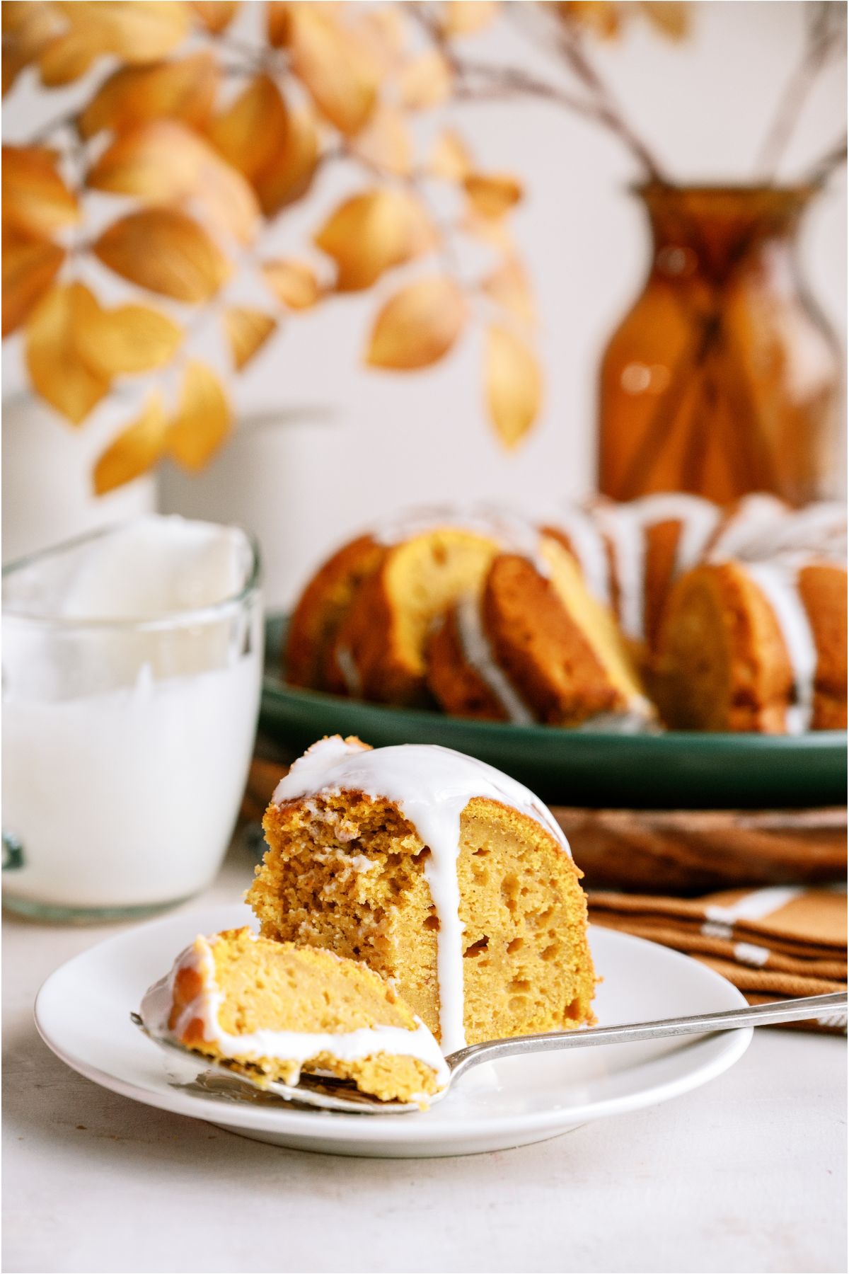 A slice of Pumpkin Bundt Cake with cream cheese glaze on a plate with a fork. The remaining Pumpkin Bundt Cake in the background.