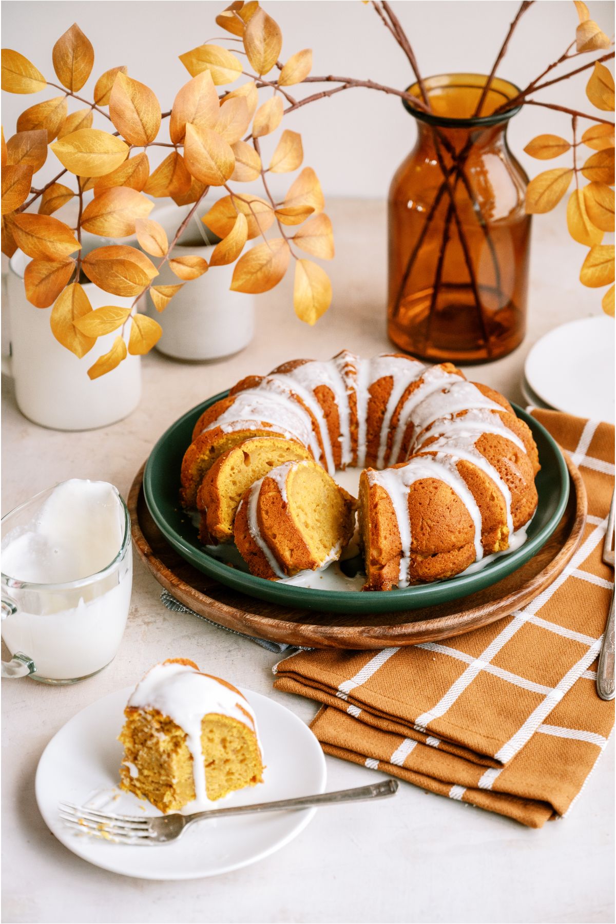 Pumpkin Bundt Cake with cream cheese frosting on a serving plate. A vase with Fall leaves and a checkered towel in the background.