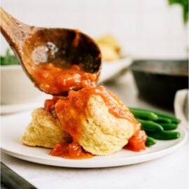 A wooden ladle placing Tomato Gravy (Traditional Southern Recipe) over two biscuits on a white plate. Green beans on the side of the plate.