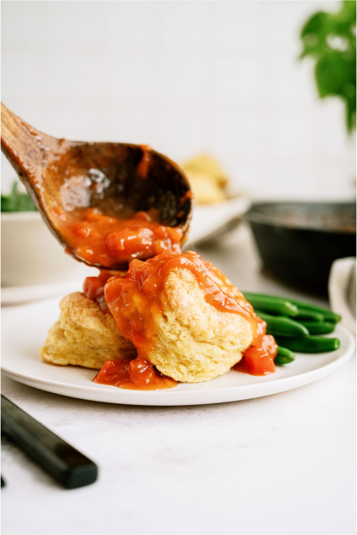 A wooden ladle placing Tomato Gravy (Traditional Southern Recipe) over two biscuits on a white plate. Green beans on the side of the plate.