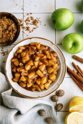 A bowl of Apple Pie Filling surrounded by green apples, cinnamon and brown sugar.