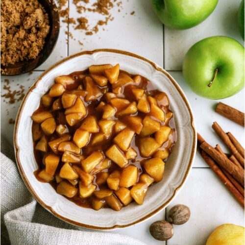 A bowl of Apple Pie Filling surrounded by green apples, cinnamon and brown sugar.