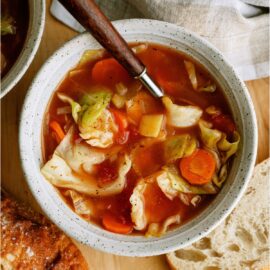 A white bowl filled with Cabbage Soup with a spoon. Surrounded by slices of crusty bread.