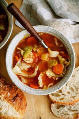 A white bowl filled with Cabbage Soup with a spoon. Surrounded by slices of crusty bread.