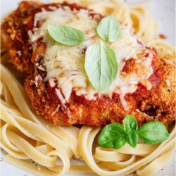 A white plate with Chicken Parmesan topped with sauce and fresh herbs served over noodles.