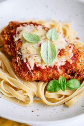 A white plate with Chicken Parmesan topped with sauce and fresh herbs served over noodles.