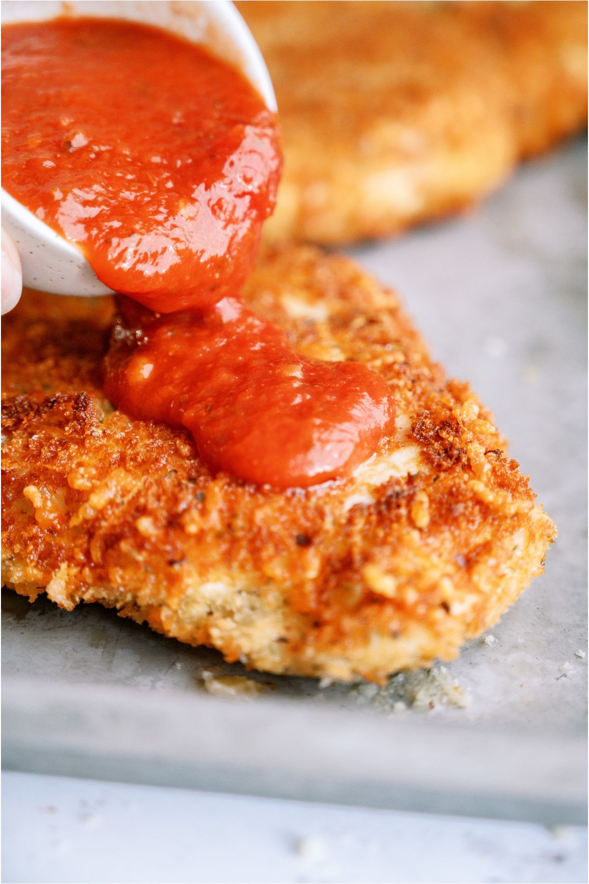 Fried panko chicken breast on a baking sheet with marinara sauce being poured on top.