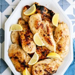 A plate full of cooked chicken breasts with slices of fresh lemons.