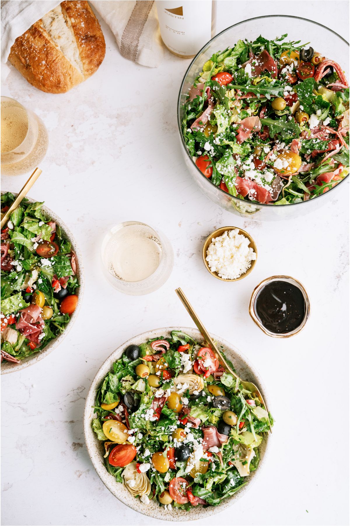 Top view of 3 bowls of Antipasto Salad with a loaf of bread and glasses surrounding the bowls.