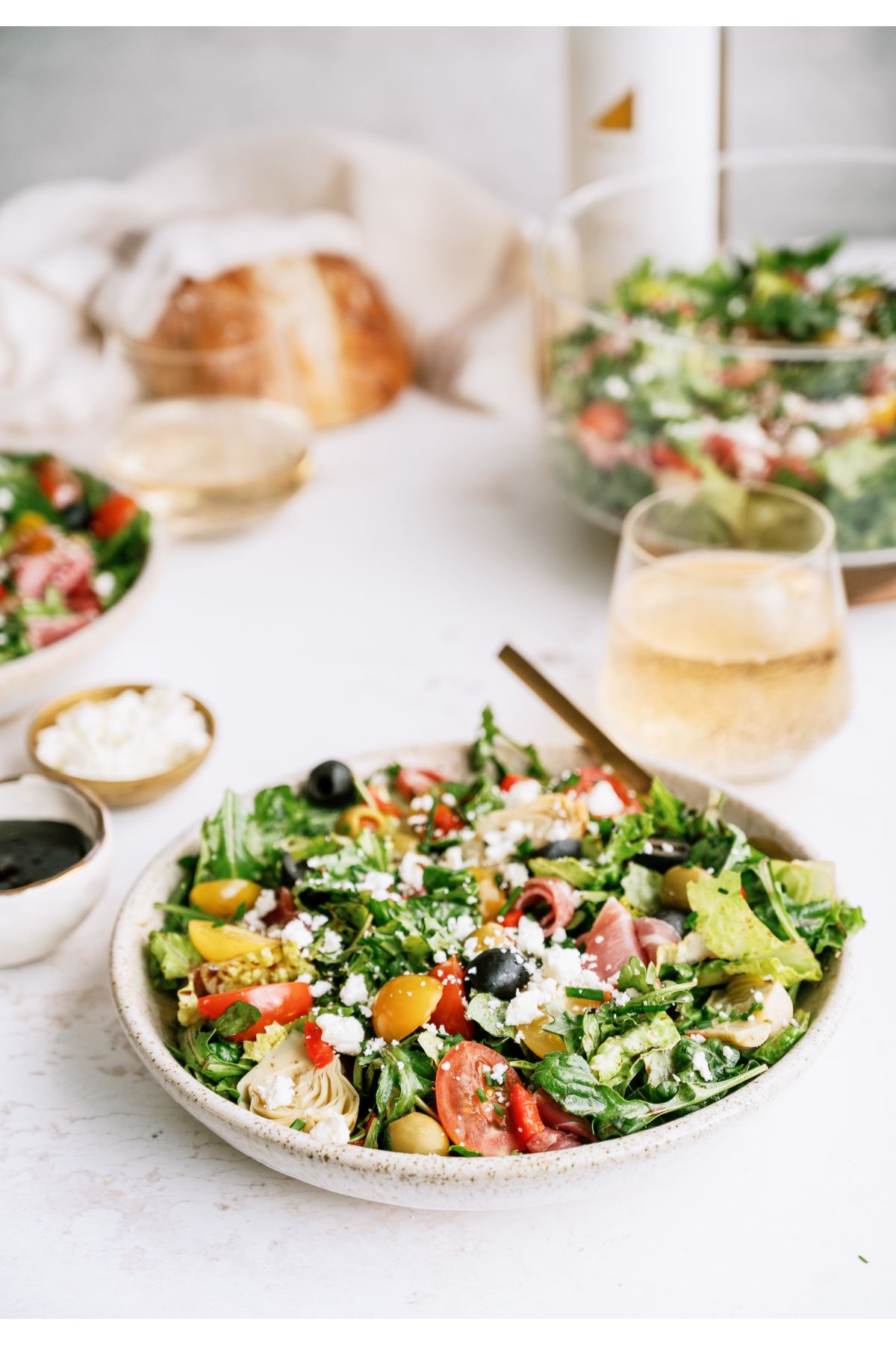 A bowl of Antipasto Salad with a fork, a glass with clear beverage and a loaf of bread in the background.