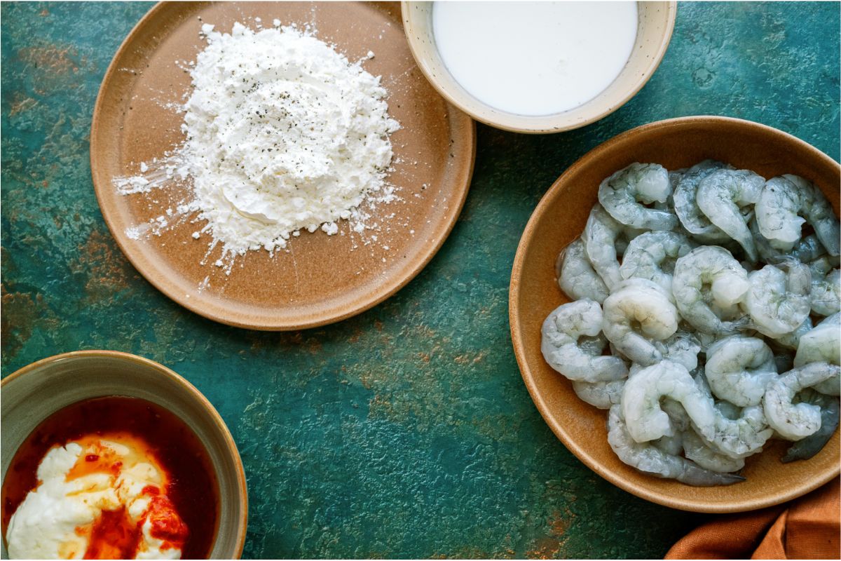 A bowl of raw shrimp, a small bowl of buttermilk, a bowl of cornstarch and a small bowl of dip.