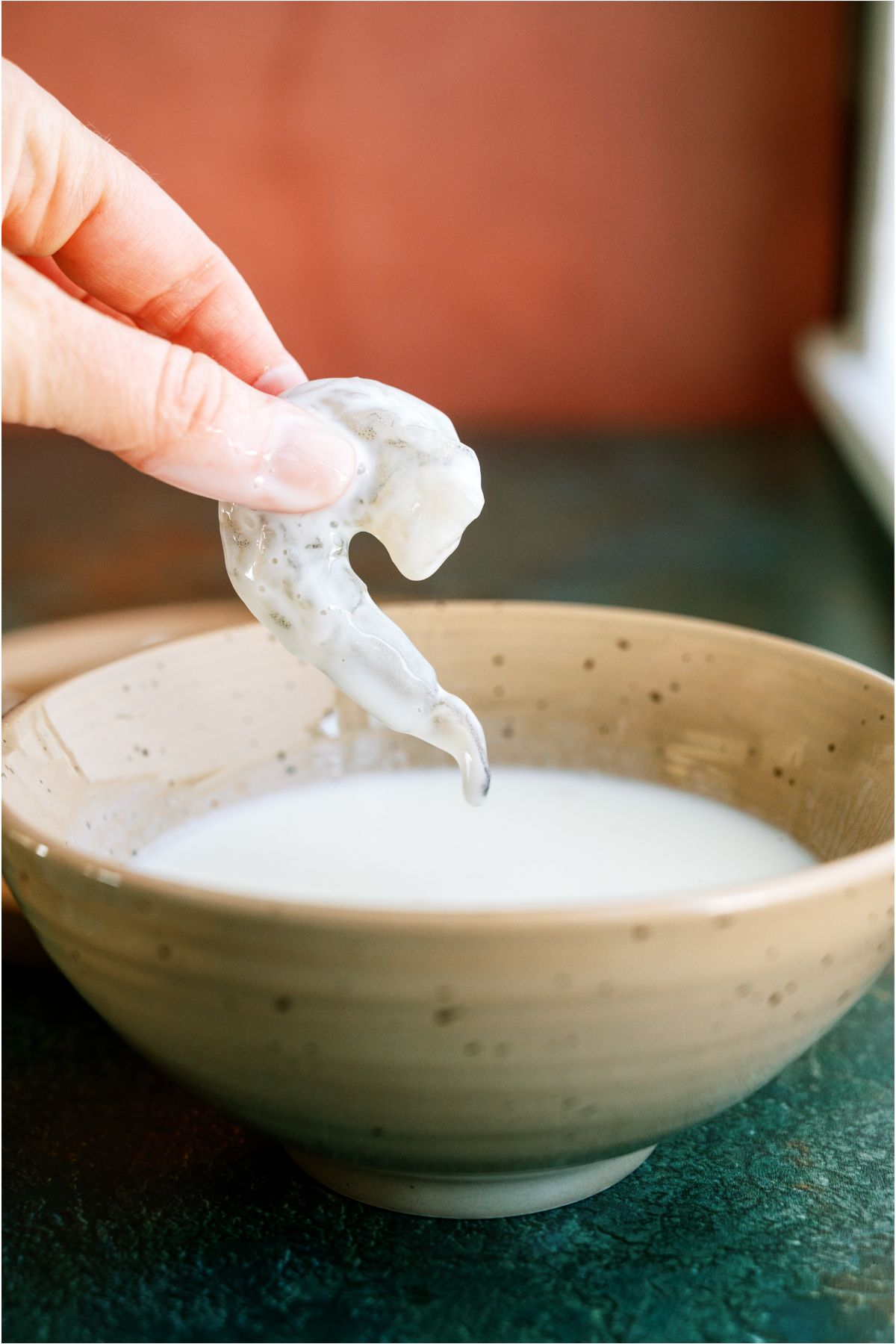 A hand dipping a raw shrimp in a shallow bowl of buttermilk.