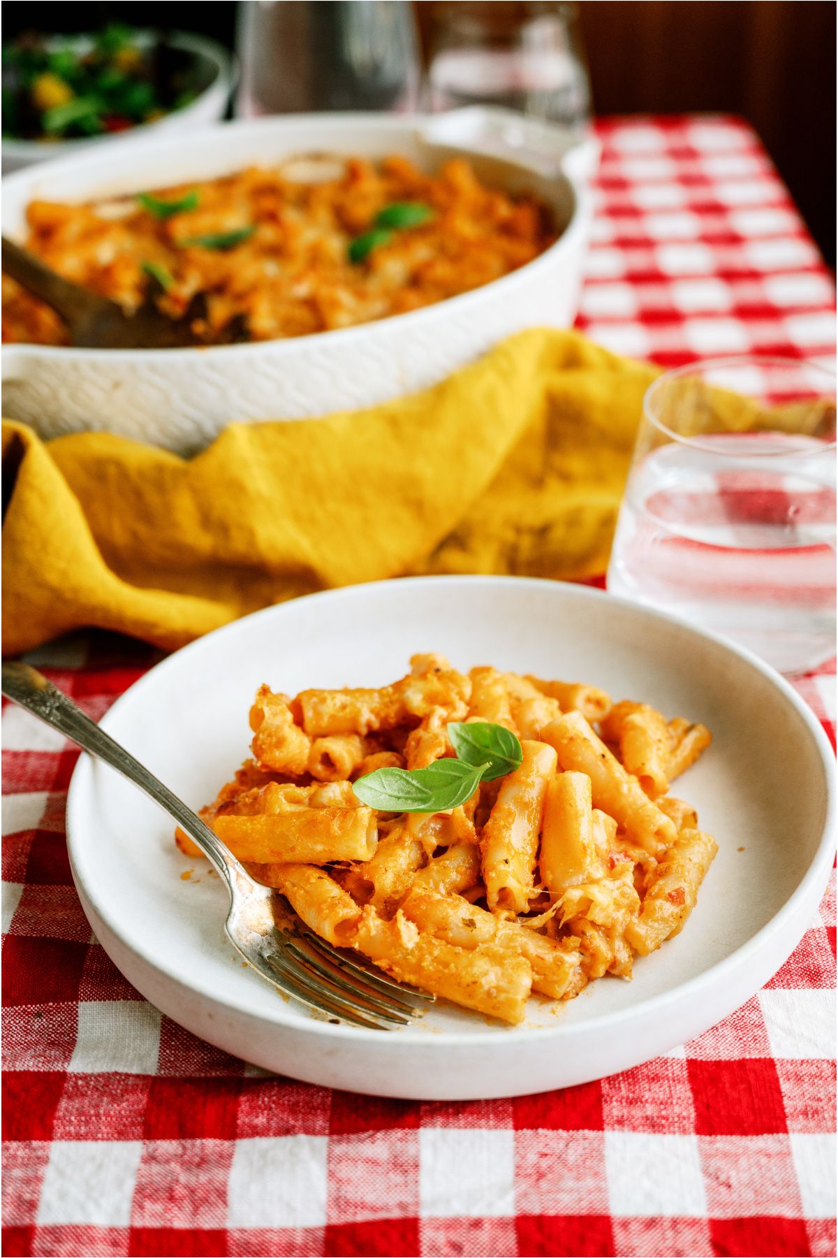 A serving of Five Cheese Ziti Al Forno on a plate with a fork. The remaining Five Cheese Ziti Al Forno in a baking dish in the background.