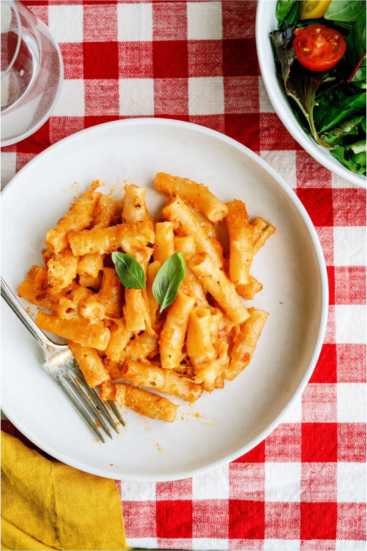 Top view of a serving of Five Cheese Ziti Al Forno on a plate with a fork. A salad bowl off to the side. A red and white checkered tablecloth as the backdrop.