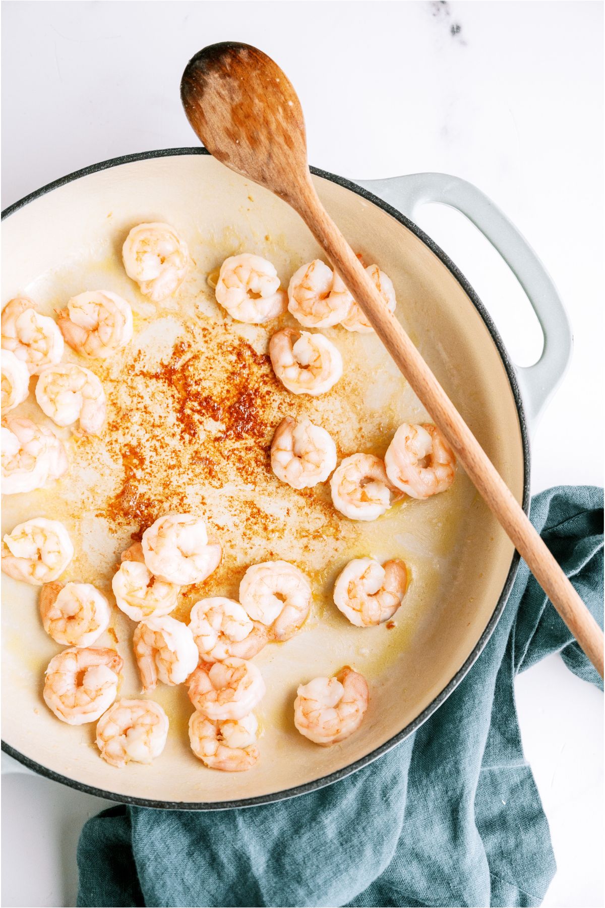 Raw shrimp being cooked in a white skillet with a wooden spoon.