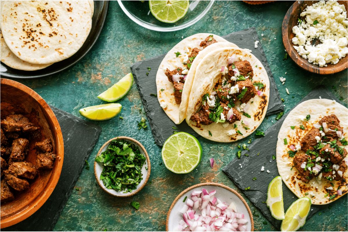 2 Street Tacos (Carne Asada) surrounded by lime wedges, a bowl of cilantro and a bowl of diced onion. Other Street Tacos (Carne Asada) off to the side.