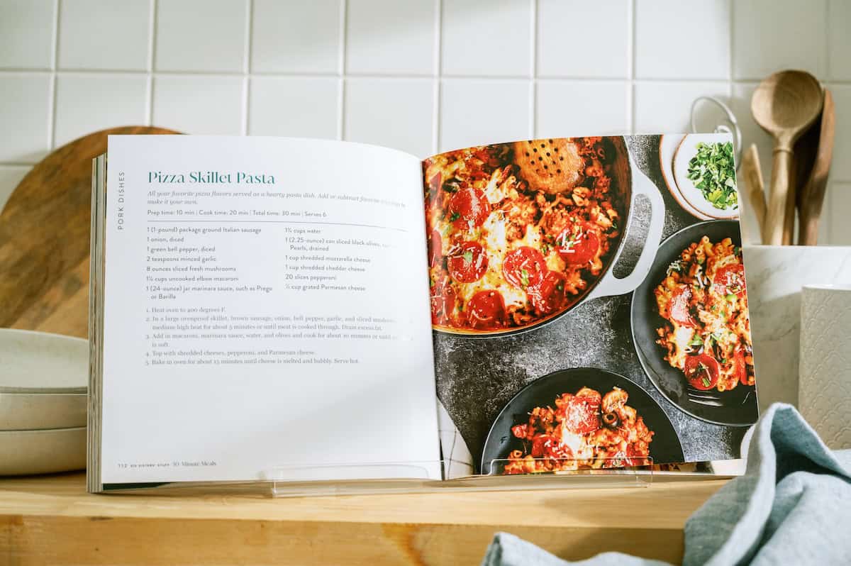 An open cookbook on a kitchen counter displays a Pizza Skillet Pasta recipe on the left and a photograph of the finished dish in a skillet on the right.